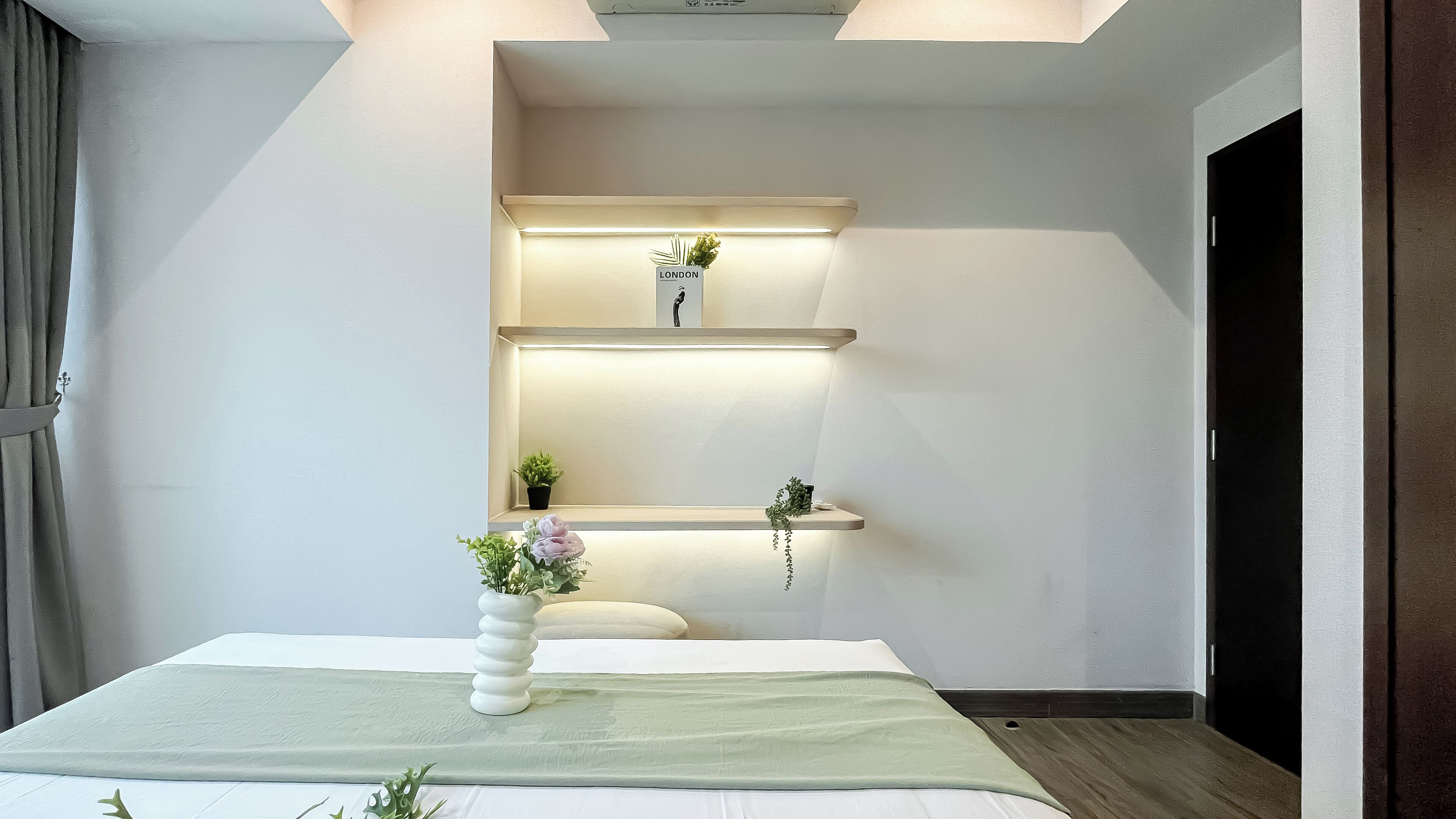 A modern hotel room with a bed, a bedside table with a vase of flowers, and two illuminated shelves with potted plants.