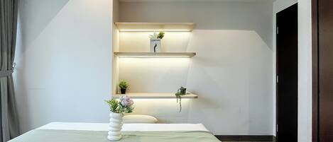 A modern hotel room with a bed, a bedside table with a vase of flowers, and two illuminated shelves with potted plants.