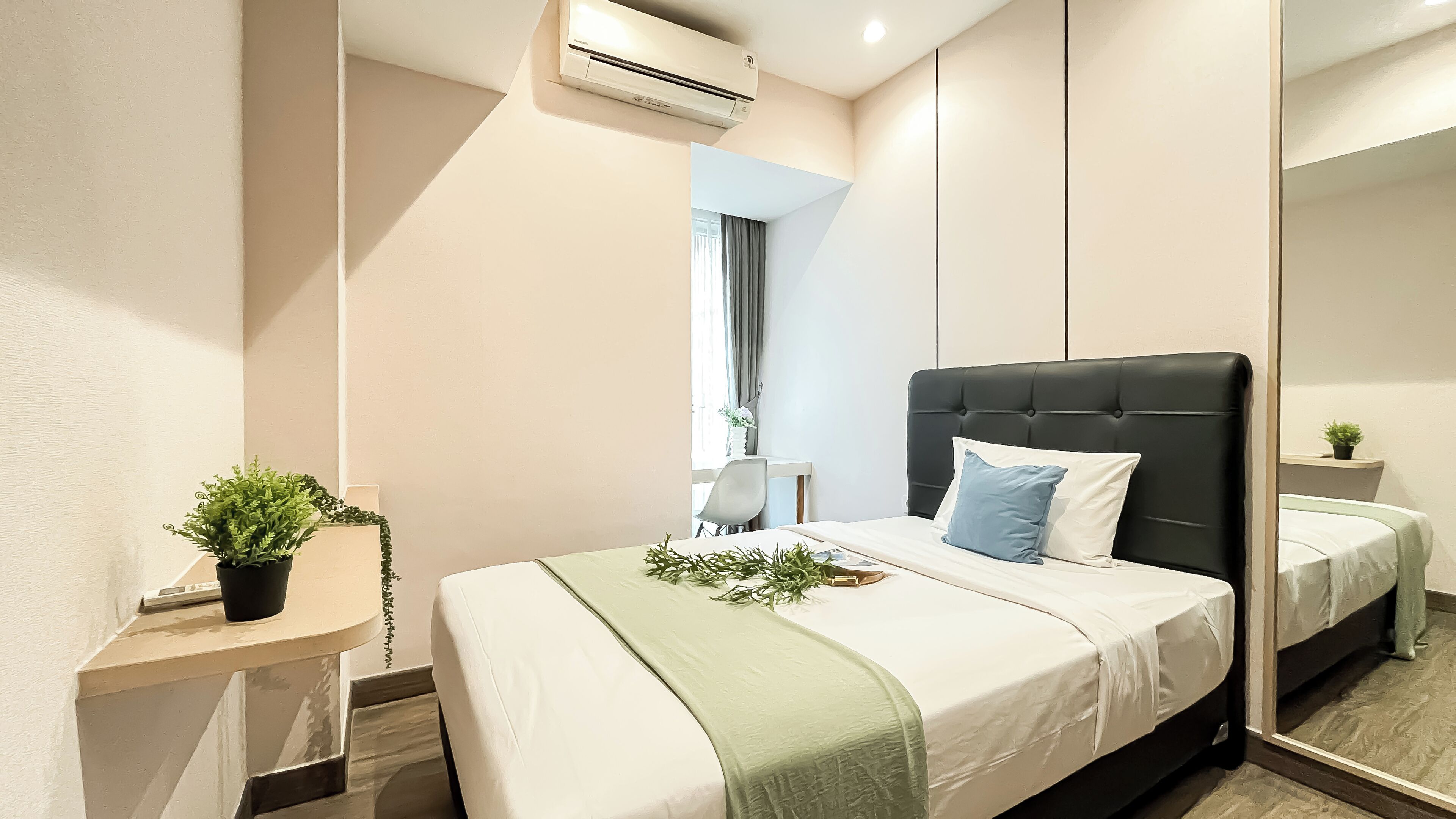A modern hotel room with a large bed, a bedside table with a plant, a small desk, and a large mirror.