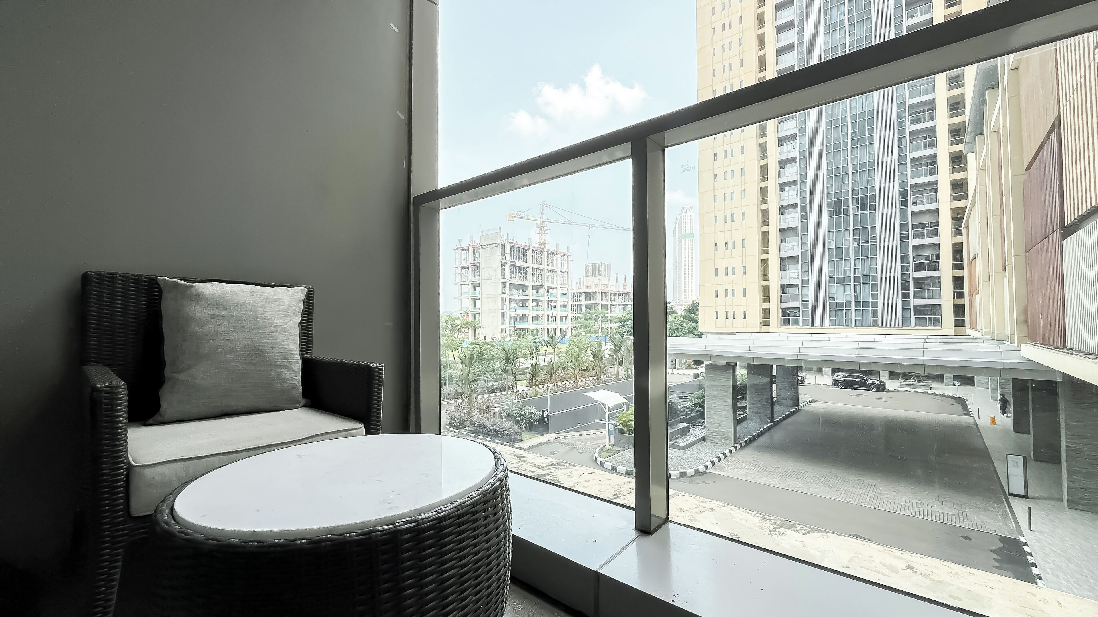 A balcony with a wicker chair and a small table, overlooking a construction site and high-rise buildings.