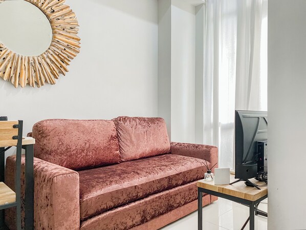 Un salon moderne avec un canapé en velours rose, une table d’appoint en bois, un téléviseur et un miroir décoratif accroché au mur.