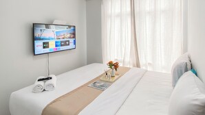 A hotel room with a bed, a TV mounted on the wall, a window with curtains, and a small table with a vase of flowers. - Good Place 1Br Vasanta Innopark Apartment (Cikarang)