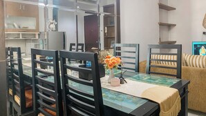 A dining area with a glass-top table, black chairs, and a vase with orange flowers. There are wooden shelves and a television on the wall. - Warm And Homey 2Br At Marina Ancol Apartment (Jakarta)