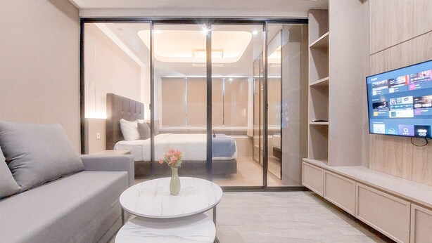 Ein modernes Hotelzimmer mit einer Couch, einem kleinen runden Tisch mit einer Vase, einem Bett mit Kopfteil, einer Schiebetür zu einem Balkon und einem an der Wand montierten Fernseher.