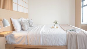 A modern bedroom with a wooden bed, white bedding, a bedside table with a lamp, and a window with natural light.