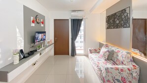 Uma sala de estar moderna com um sofá, uma televisão de tela plana na parede, uma mesinha com um vaso e uma porta que dá para outro quarto.