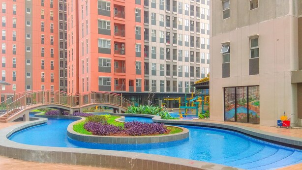 Piscina com uma ponte, rodeada por vegetação e um edifício com várias andares e varandas.