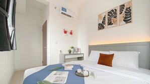 Cama bem arrumada com almofadas brancas e castanhas, um cobertor azul e uma bandeja com livros e uma planta. Há um ar condicionado fixado na parede e uma obra de arte acima da cama.