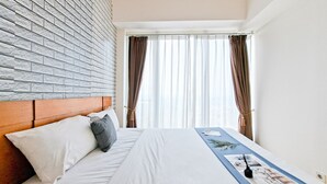 Une chambre d’hôtel avec une tête de lit décorée d’un motif en briques, un lit bien fait avec une literie blanche et grise, une table de chevet bleue sur laquelle se trouvent un magazine et une télécommande, et une fenêtre ornée de rideaux légers.