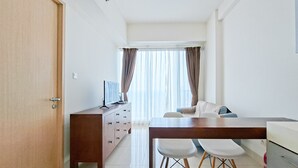 A modern, well-lit room with a wooden dining table, white chairs, a sofa, and a TV on a wooden cabinet. - Simple Look 1Br Apartment At Tree Park City Bsd (Tangerang)