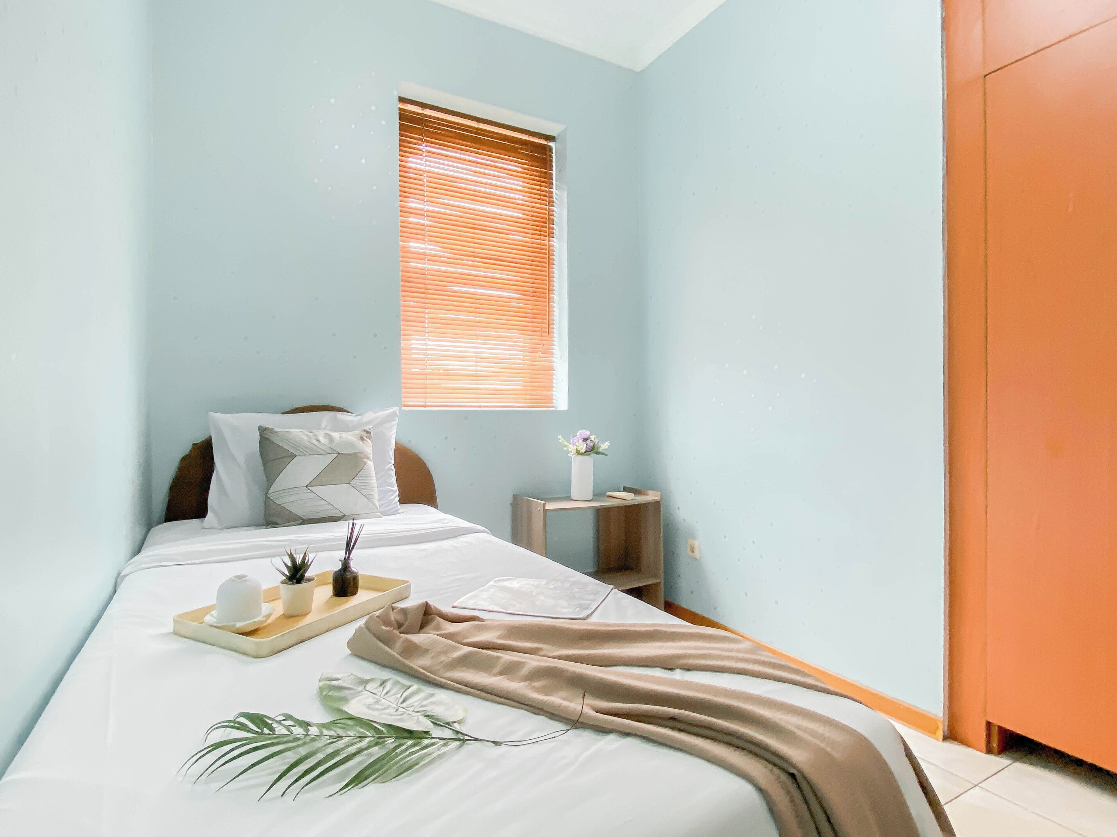 Ein ordentlich gemachtes Bett mit einer beigen Decke, ein Nachttisch mit Blumen in einer Vase und ein Fenster mit Jalousien.