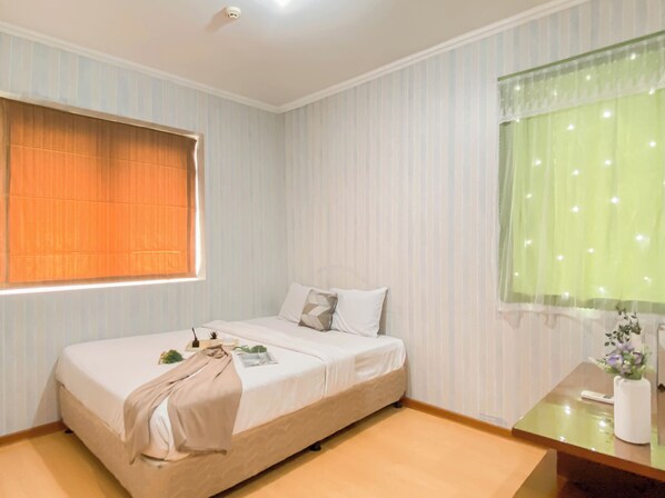 A single bed with a beige blanket, a bedside table with a vase of flowers, and a window with orange blinds. - Compact And Functional 3Br Apartment Grand Palace Kemayoran (Jakarta)
