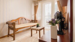 A living room with a wooden sofa, a small table with a vase of flowers, and a standing fan. - Compact And Functional 3Br Apartment Grand Palace Kemayoran (Jakarta)
