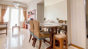 A dining area with a wooden table, chairs, and a stool. There is a small table with a plant and a shelf with decorative items. - Compact And Functional 3Br Apartment Grand Palace Kemayoran (Jakarta)