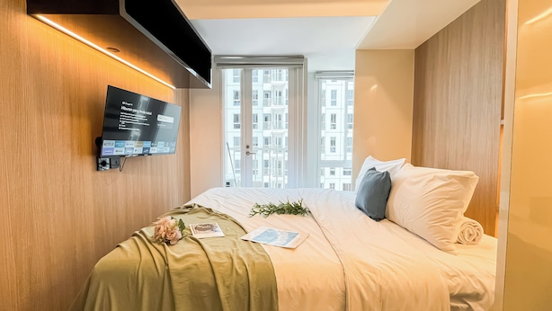Ein modernes Hotelzimmer mit einem großen Bett, einem Wand-Fernseher und Blick auf die Stadt durch große Fenster.