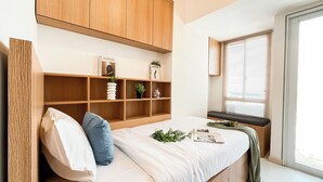 A bedroom with a bed, a wooden shelf unit, a window with blinds, and a small table with a plant.