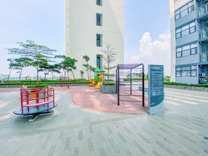 Ein Spielplatz mit Rutschen und Klettergerüsten, umgeben von einem befestigten Bereich mit Bänken und einem Hinweisschild.
