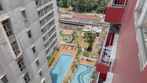 A high-rise building with multiple balconies, a swimming pool with two sections, and a red building in the background. - Nice & Cozy Studio Apartment At Taman Melati Margonda (Depok)