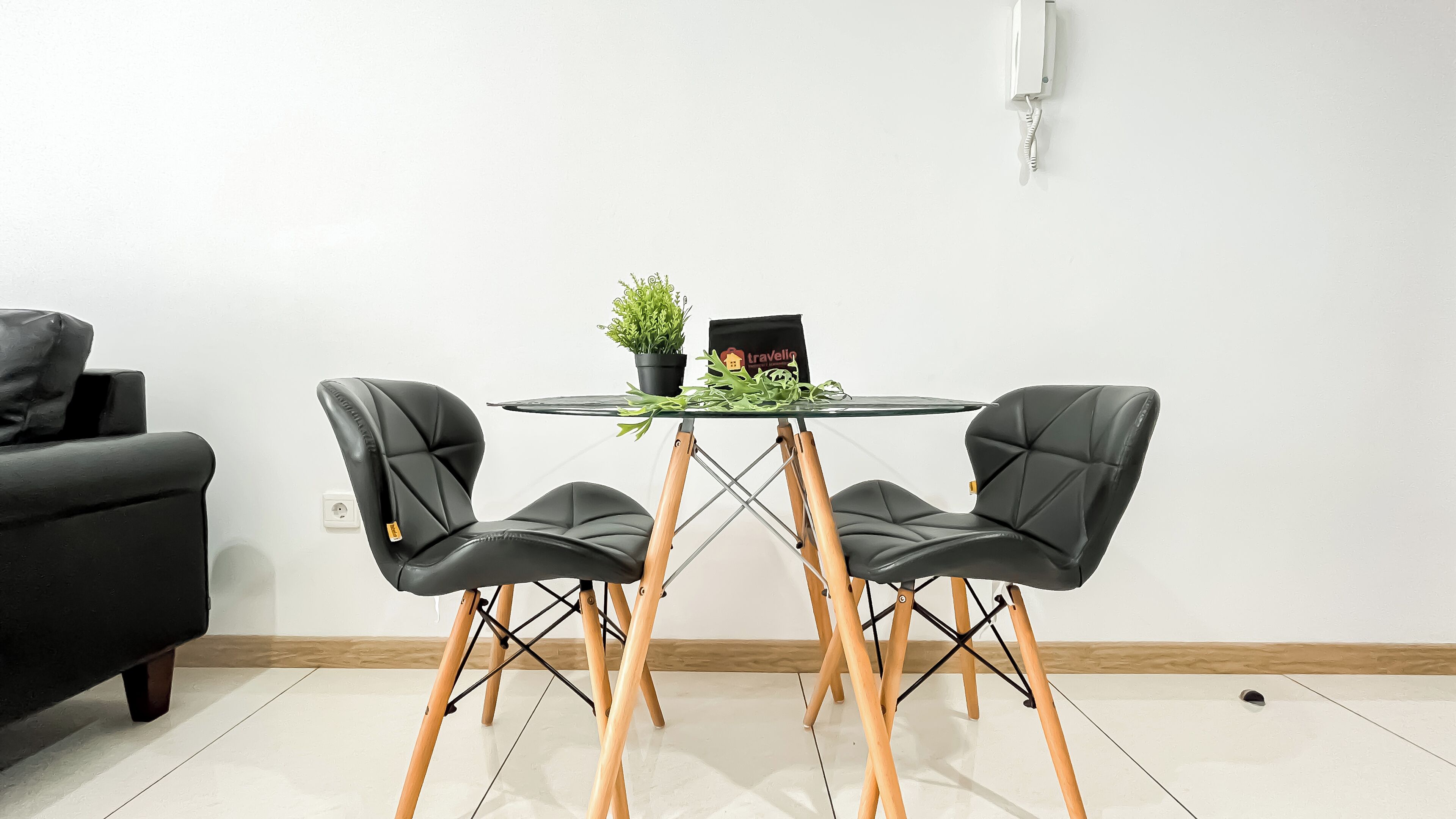Una habitación moderna con una mesa redonda de vidrio, dos sillones de cuero negro, una pequeña planta en maceta y una bolsa de la marca 'Travelly'.