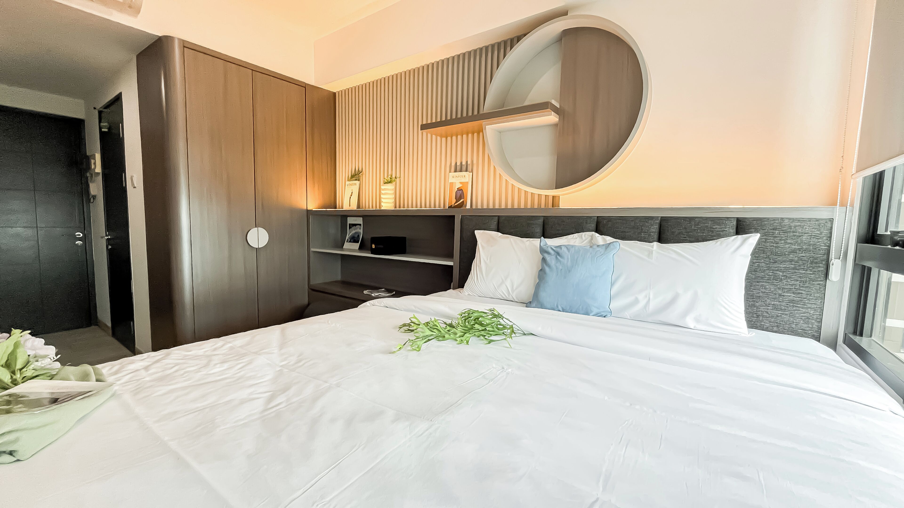 A modern hotel room with a large bed, a wardrobe, a round mirror, and a bedside table.