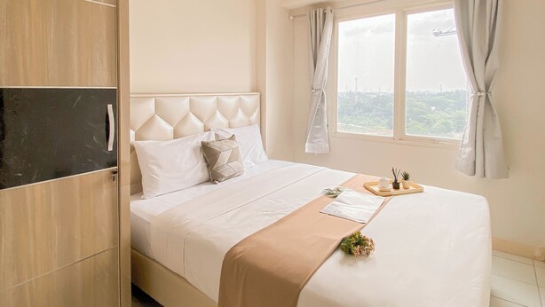 A neatly made bed with a headboard, a bedside table with a tray, and a window with curtains. - Simple Look 2Br At Apartment Podomoro Golf View (Gunung Putri)