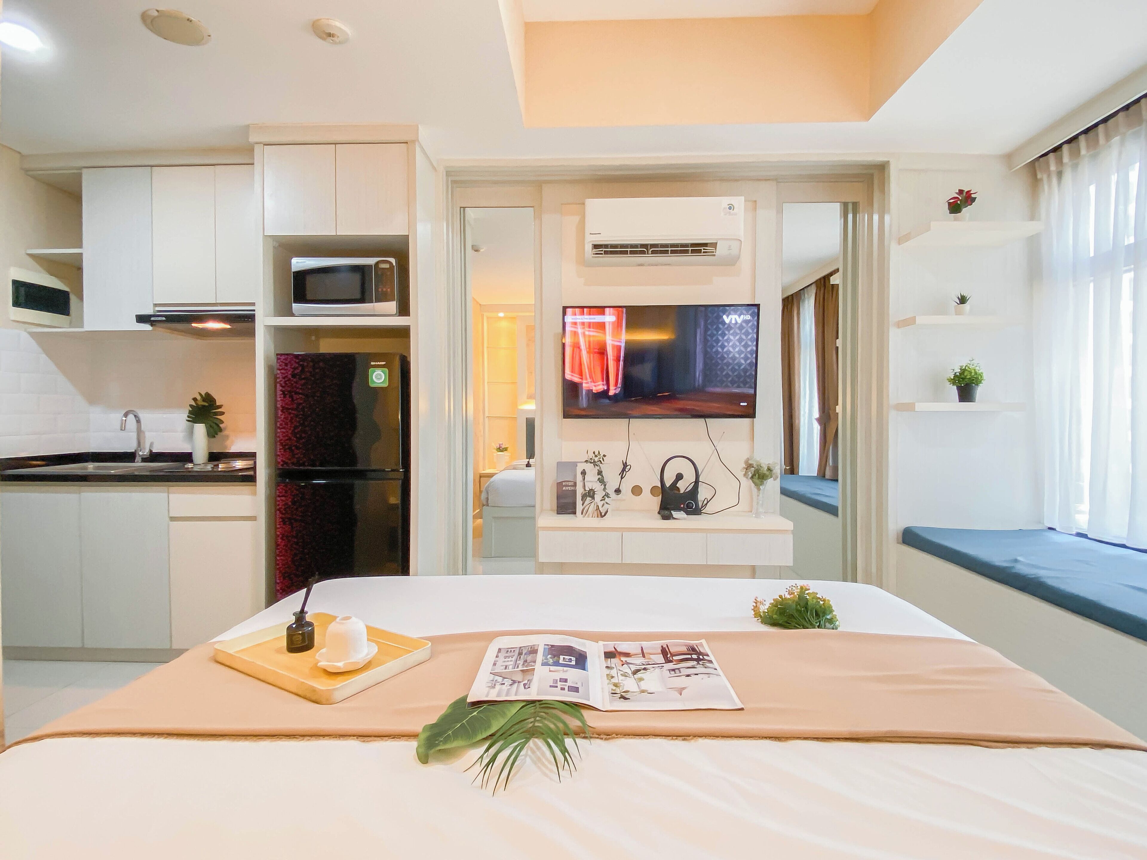 Habitación de hotel moderna con kitchenette, una cama con una revista y una planta, televisión y ventana con cortinas.