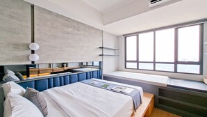 A modern hotel room with a large bed, built-in bench seating, a window with a city view, and a concrete accent wall. - 1Br Near Alam Sutera Mall At Collins Boulevard Apartment (Tangerang)