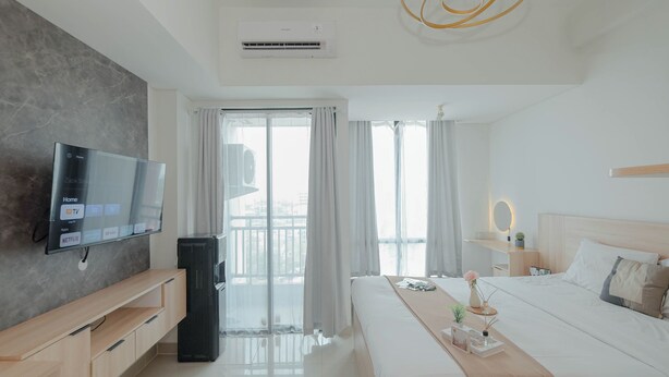 A modern hotel room with a flat-screen TV, a large bed, a bedside table with a lamp, and a view of the city through the window. - Minimalist Studio At Apartment Lrt City Bekasi (Bekasi)