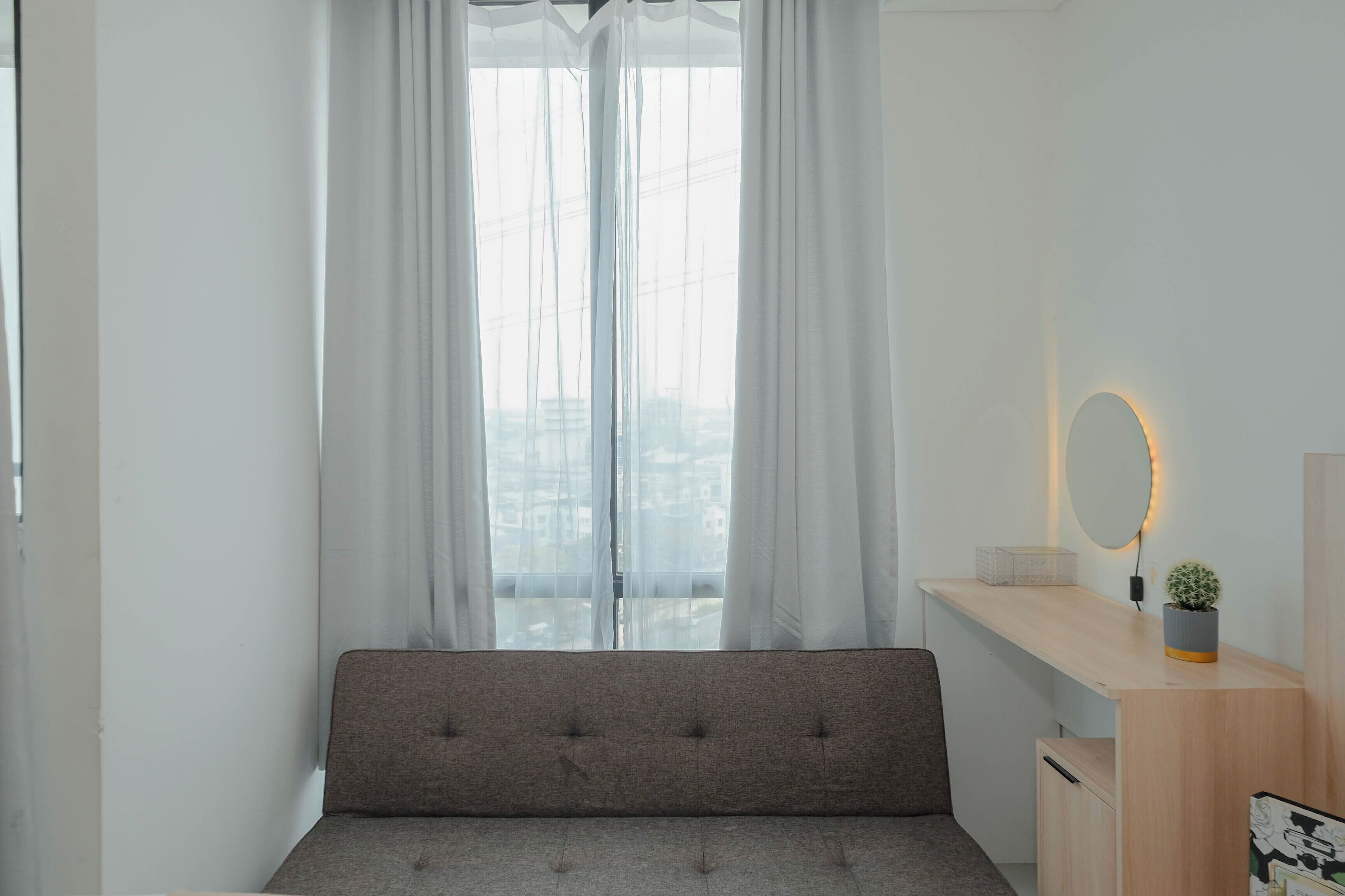 A bedroom with a bed, a bedside table with a mirror, a small plant, and a window with sheer curtains.