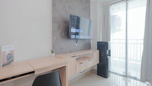 A modern hotel room with a flat-screen TV mounted on the wall, a wooden desk with a chair, a small potted plant, and a water dispenser. - Minimalist Studio At Apartment Lrt City Bekasi (Bekasi)
