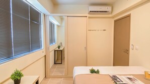 A hotel room with a bed, a bedside table, a small plant, a window with blinds, a door, and an air conditioning unit. - Warm Neutral 2Br At Meikarta Apartment (Cikarang)