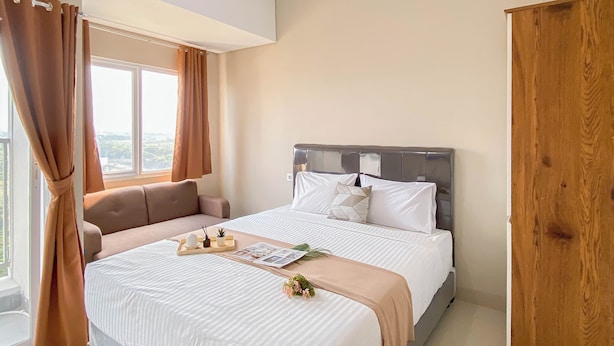 A bedroom with a bed, a window with curtains, a bedside table with a tray, and a wooden wardrobe. - Neutral Minimalist Studio Apartment At Lrt City Sentul - Royal Sentul Park (Bogor)