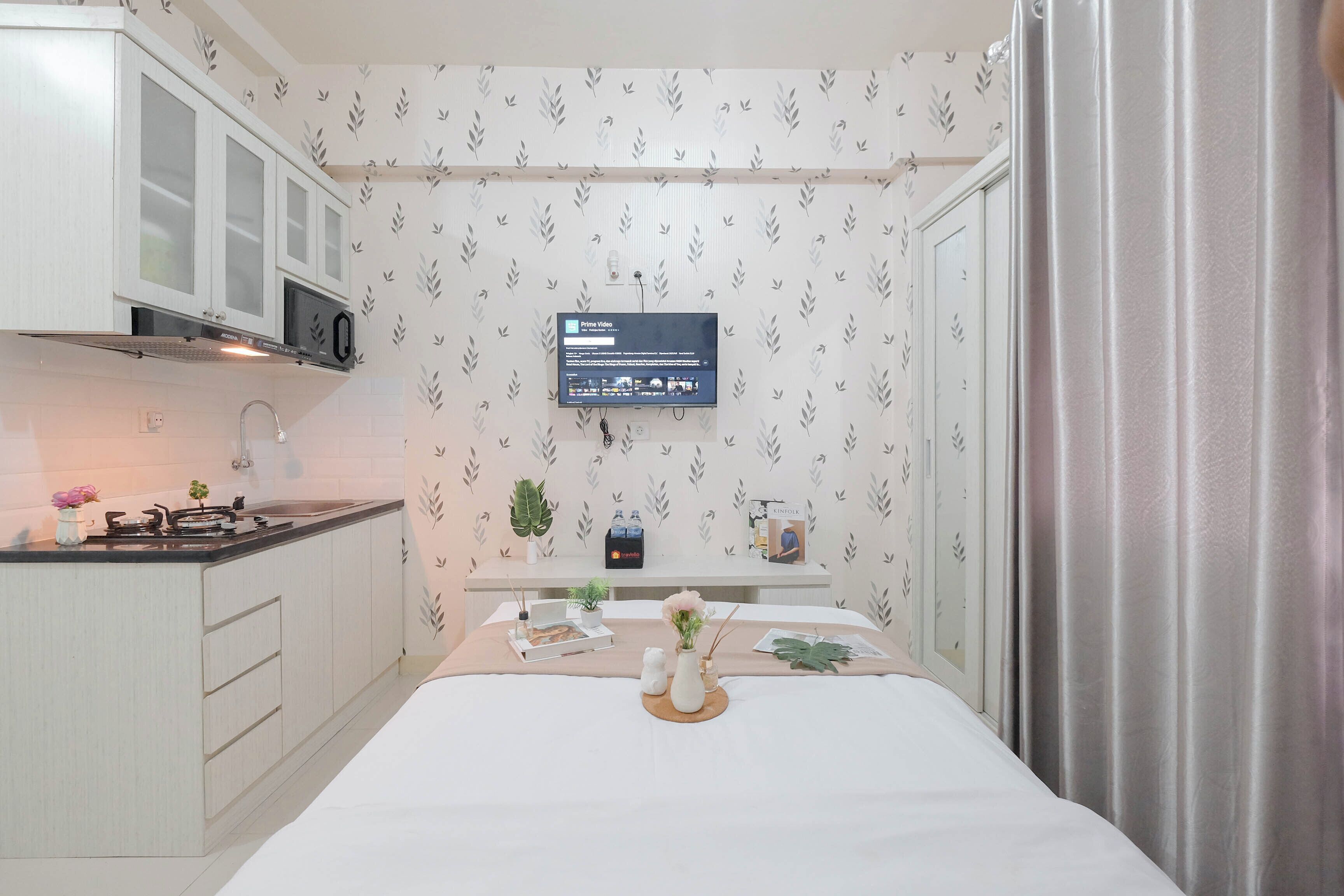 A compact hotel room with a kitchenette, a flat-screen TV mounted on the wall, and a bed with a white bedspread.