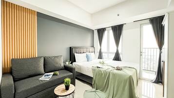 A modern hotel room with a grey sofa, a bed with white linens, a green table with a plant, and a view of the outdoors through large windows.