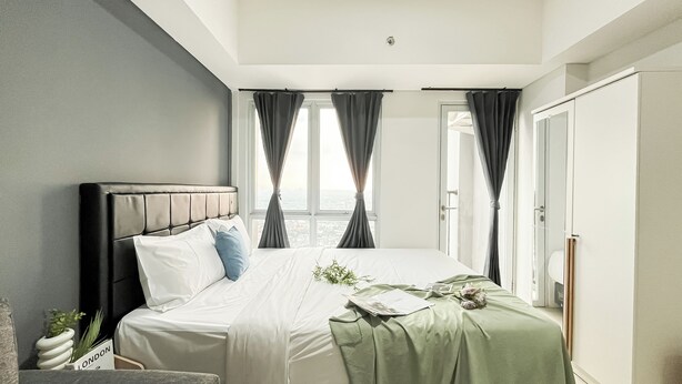 A modern bedroom with a large bed, grey curtains, and a view of the city. - Good Deal Studio At Apartment Tamansari Bintaro Mansion (South Tangerang)