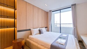 A modern hotel room with a large bed, wooden panel walls, a small wooden stool, and a view of the city through a large window. - Cozy Warm Studio Apartment At Collins Boulevard (Tangerang)