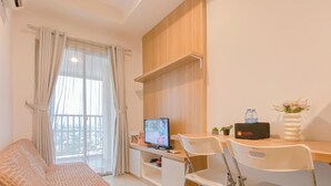 A hotel room with a bed, a desk with a computer, two chairs, a TV, a window with curtains, and a small potted plant. - Comfortable 1Br At Belmont Residence Puri Apartment (Jakarta)