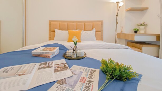 A neatly made bed with a blue bedspread, a tray with books and a vase of flowers, and a lamp on a nightstand. - Comfortable 1Br At Belmont Residence Puri Apartment (Jakarta)