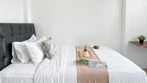A neatly made bed with white linens and a beige throw blanket, a bedside table with a small plant and a tray, and a dark upholstered headboard. - Studio Apartment Jkt Living Star Near Citimall Cimanggis (Jakarta)