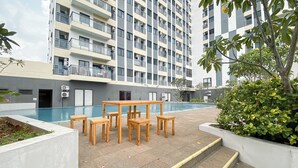A poolside area with a wooden table and chairs, surrounded by greenery and a multi-story building with balconies. - Studio Apartment Jkt Living Star Near Citimall Cimanggis (Jakarta)