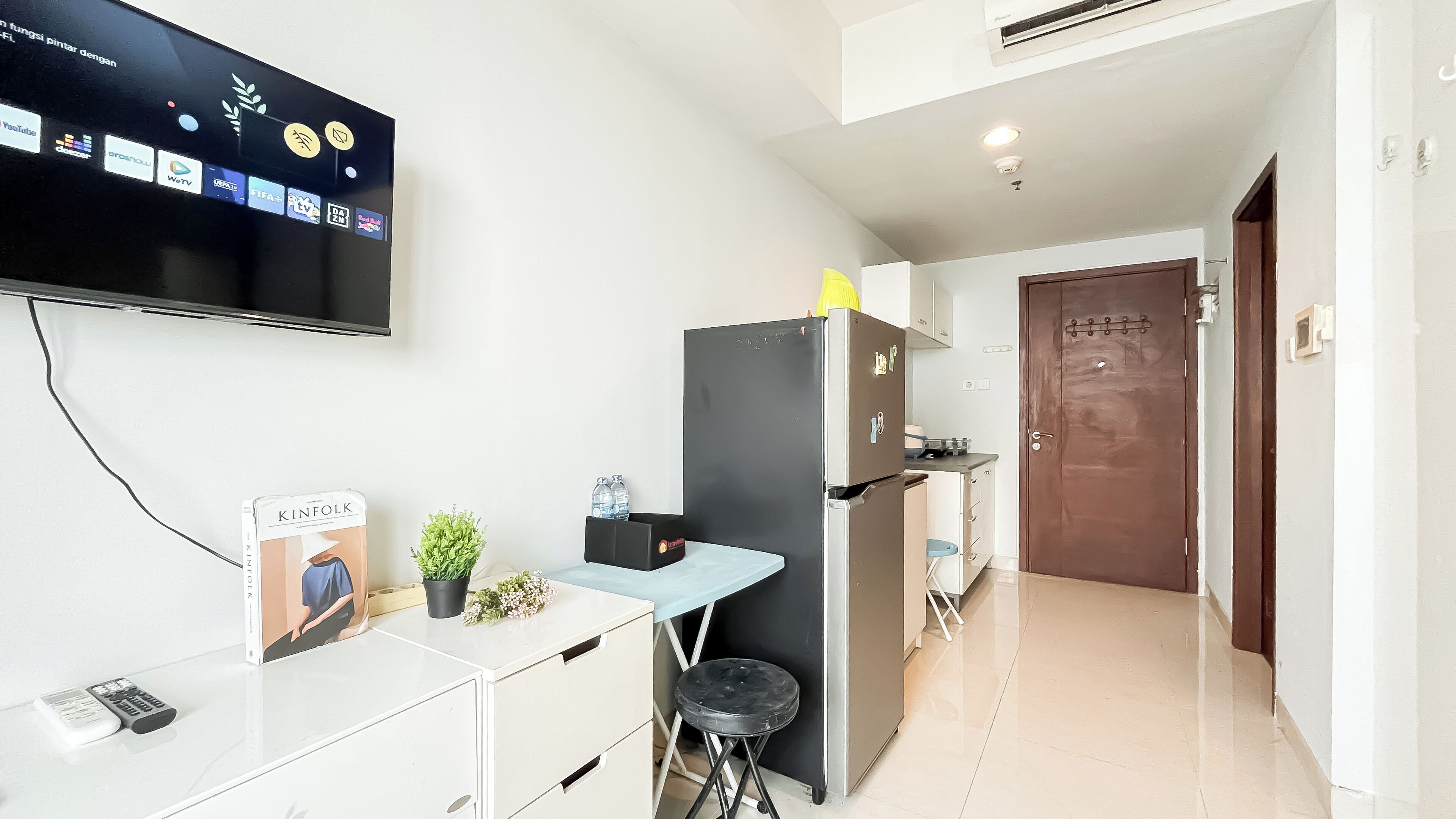 A compact kitchen with a refrigerator, a small dining table, and a TV mounted on the wall displaying various apps.