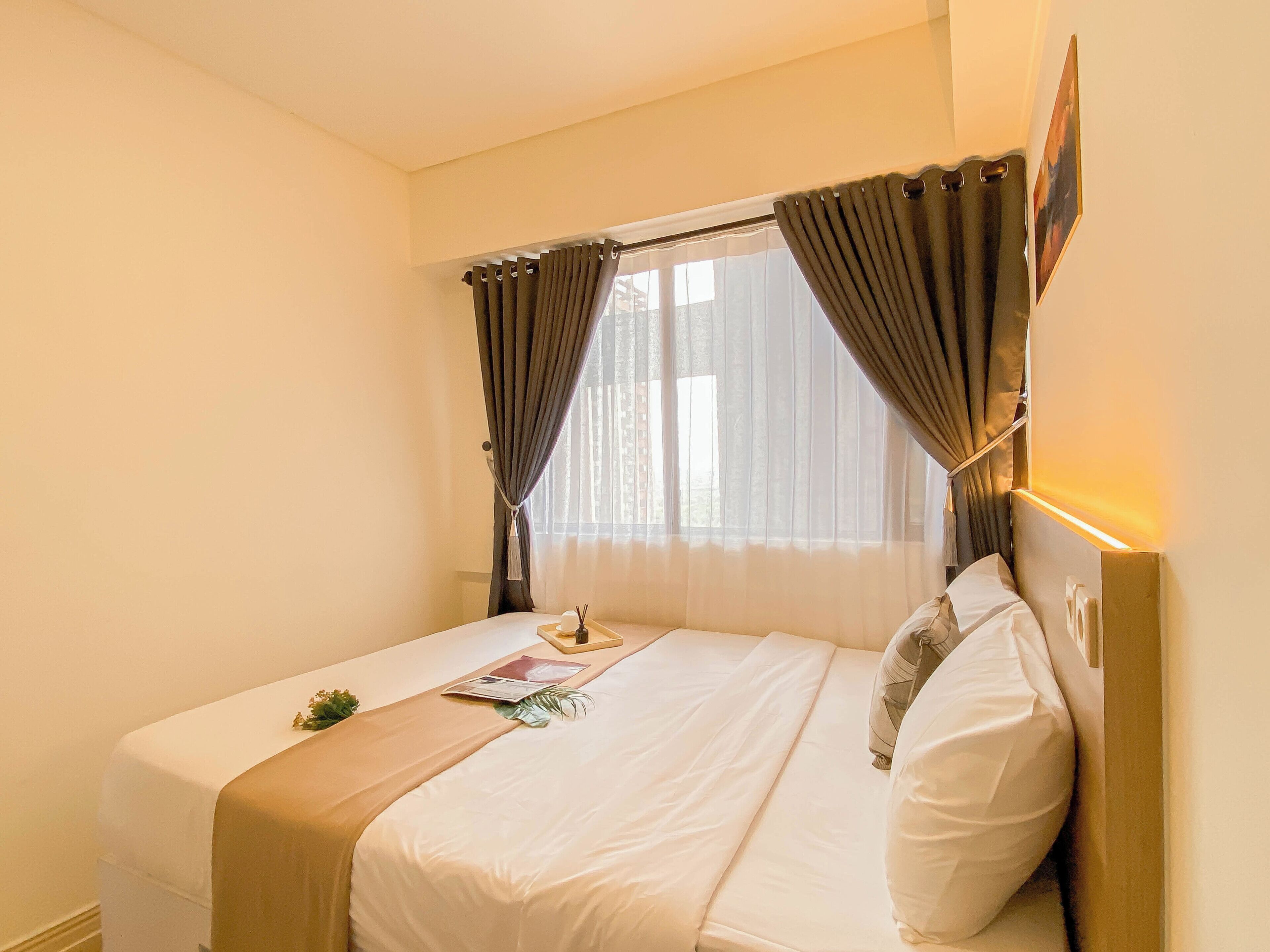 Una cama bien hecha con edredón blanco y almohadas, una mesita de noche con lámpara y una ventana con cortinas.