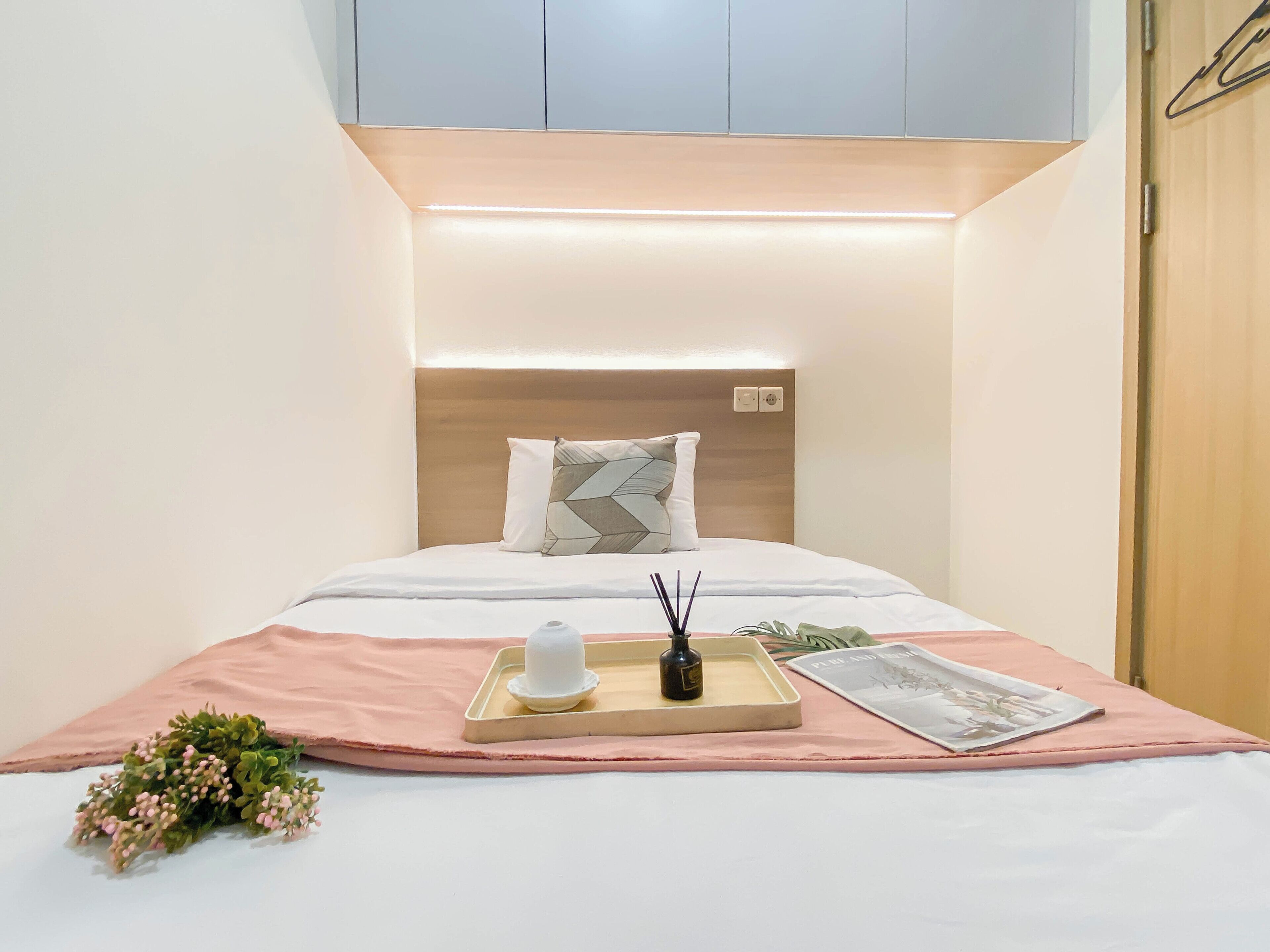 A neatly made bed with a headboard, a tray with a bottle and a candle, and a small bouquet of flowers on a pink cloth.