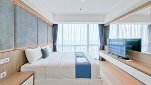 A modern hotel room with a large bed, a flat-screen TV, and a view of the city through large windows. - Modern And Spacious 3Br At Apartment Hillcrest House (Curug)