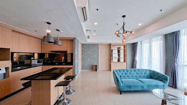 A modern kitchen with wooden cabinets, a black countertop, and stainless steel appliances. There is a blue sofa and a glass coffee table in the living area. - Modern And Spacious 3Br At Apartment Hillcrest House (Curug)
