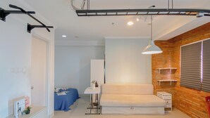 A bedroom with a white bed, a desk with a chair, a small table with a lamp, and a window with blinds. - Spacious Studio At Signature Park Grande Apartment (Jakarta)
