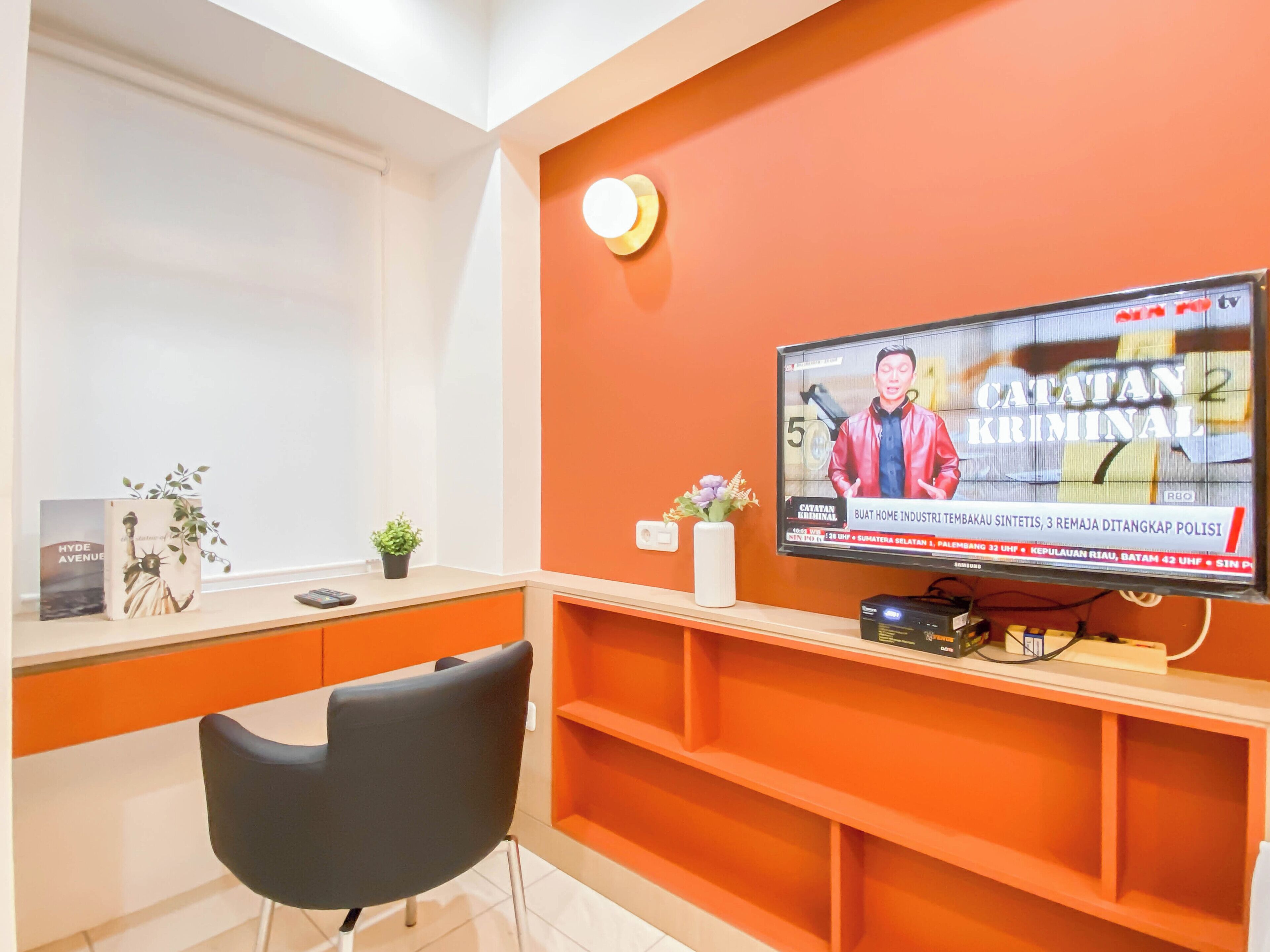 A room with a television displaying a news program, a desk with a plant, a chair, and a shelf with a small decorative item.