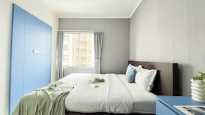 A hotel room with a bed, a nightstand, a window with curtains, and a view of buildings outside. - Cozy And Modern 2Br Apartment Marina Ancol (Jakarta)