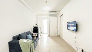A living room with a dark-colored sofa, a flat-screen TV mounted on the wall, and a hallway leading to a door. - Basic 2Br Apartment At Green Bay Pluit (Jakarta)
