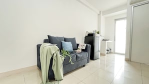 A modern living room with a dark grey sofa, a small black refrigerator, and a white kitchen area. - Basic 2Br Apartment At Green Bay Pluit (Jakarta)